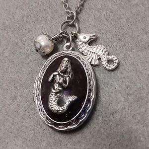 Puede incluir: Collar con colgante de sirena plateada con un dije de caballito de mar y un acento de perla. El colgante ovalado presenta un diseño de sirena sobre un fondo oscuro. El collar está sobre un fondo gris.
