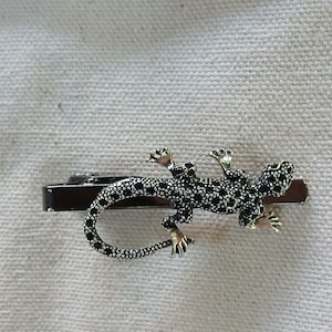 May include: A silver tie clip featuring a black and white lizard design. The lizard is detailed with black spots and has a gold-toned finish.