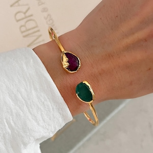 Könnte beinhalten: Ein goldfarbenes Armband in offener Form mit zwei asymmetrischen, gefassten Steinen. Ein Stein ist dunkelrot und tropfenförmig, der andere ist kräftig grün und unregelmäßig geformt. Das Metallband hat eine glatte, polierte Oberfläche.