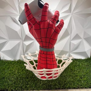 May include: A red hand sculpture with black web patterns holding a white gaming controller. The hand features a grey wrist band and emerges from a white, circular, web-like base. The design mimics a superhero aesthetic with a vibrant red finish.