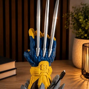 May include: A blue and yellow glove-shaped stand featuring three long, silver, pointed blades extending upward from the knuckles. The base is designed to look like jagged gray rocks and includes a black USB-C charging cable extending from the side.