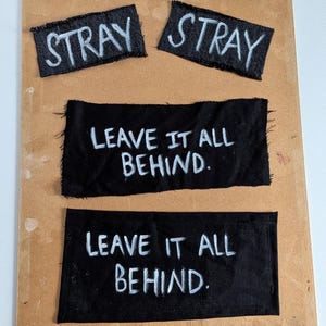 May include: Four rectangular black fabric patches with frayed edges. Two smaller patches feature the white text STRAY. Two larger patches feature the white text LEAVE IT ALL BEHIND. The patches are arranged in two rows on a flat tan surface.