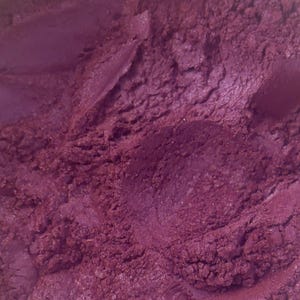 May include: A fine, loose powder pigment in a deep, rich magenta-purple hue. The texture appears granular and slightly clumped, with a subtle metallic or pearlescent sheen that catches the light across the uneven, textured surface of the powder.