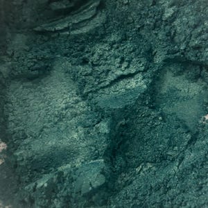 May include: A loose, finely milled cosmetic powder in a deep, metallic teal green shade. The texture appears dense and slightly clumped, showcasing a shimmering, reflective finish across the uneven surface of the pigment particles.