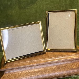 May include: Two rectangular picture frames with thin, polished gold-tone metal borders. One frame is positioned horizontally, while the other is oriented vertically. Both frames feature clear glass fronts and contain plain, light tan cardboard inserts.