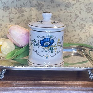 May include: A white ceramic lidded jar with a blue floral pattern and silver trim, resting on a rectangular silver-toned metal tray with four decorative feet. Two artificial tulips in shades of pink and cream are placed beside the jar.