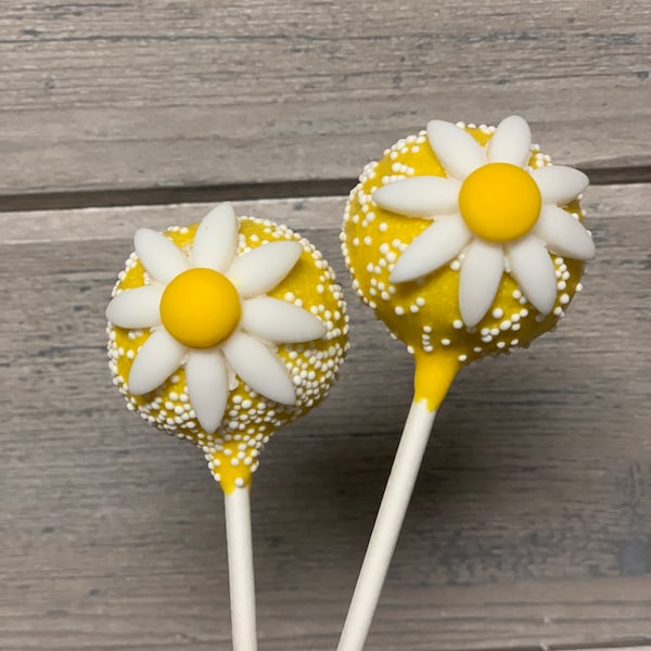 May include: Two yellow cake pops decorated with white daisy flowers and white sprinkles on white sticks.