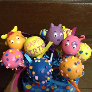 May include: A colorful assortment of cake pops decorated with sprinkles and characters. The cake pops are arranged in a blue and white polka dot container. The cake pops are decorated with the words "Happy Birthday".