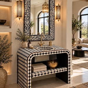 Puede incluir: Un mueble de lavabo con patrón geométrico en blanco y negro, que incluye un lavabo integrado, un grifo de latón y un estante abierto con toallas dobladas y un cuenco dorado. Arriba, un espejo rectangular a juego está flanqueado por dos apliques.