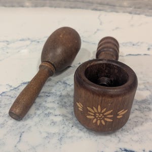 May include: A dark brown wooden nutcracker tool consisting of two separate pieces. One piece is a cylindrical base with a carved flower design on the side, and the other is a matching wooden plunger with a rounded top and a long handle.