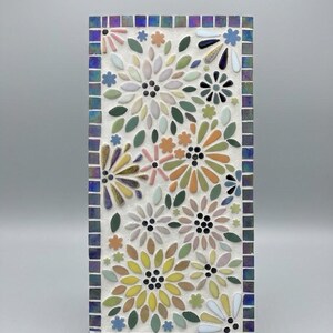 Peut inclure: Une plaque murale en mosaïque rectangulaire présentant un motif floral de pétales multicolores dans des tons de jaune, rose, vert et blanc sur fond blanc. La pièce est encadrée par une bordure de petites tuiles carrées bleu et violet irisé.