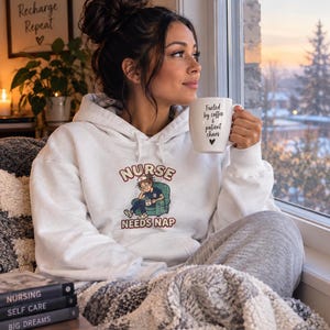 May include: A white hooded sweatshirt featuring a graphic of a nurse sitting in a chair with the text NURSE NEEDS NAP. The sweatshirt is paired with a white ceramic mug printed with the text Fueled by coffee & patient chaos and a small heart.