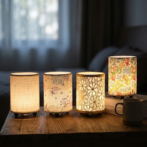 Könnte beinhalten: Vier zylindrische Tischlampen mit Stoffschirmen stehen auf einer Holzoberfläche. Eine Lampe hat eine schlichte beige Textur, eine zeigt ein blau-rosa Pusteblumenmuster, eine hat goldene Blumenumrisse und eine zeigt ein buntes Blumenmuster.