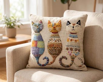 Op de afbeelding: Een vierkant beige sierkussen met drie geborduurde katten die naast elkaar zitten. Elke kat heeft een uniek, kleurrijk patroon met gedetailleerd stikwerk, lange snorharen en een gekrulde staart op een effen, lichtgekleurde achtergrond.