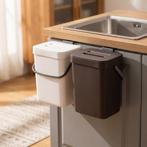 May include: Two rectangular waste bins, one white and one dark brown, are suspended from the edge of a kitchen counter. Each bin features a matching lid with a central handle slot and a side-mounted carrying handle, providing a compact storage solution.