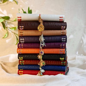 May include: A vertical stack of ten rectangular journals with textured covers in white, brown, tan, dark purple, orange, black, reddish-brown, blue, and red. Each journal features colorful stitched lacing and a central hanging metal charm.
