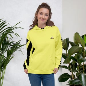 Può includere: Una felpa con cappuccio giallo brillante dotata di una tasca a marsupio frontale, strisce diagonali nere sulle maniche e un piccolo logo sul petto con il testo "Future Style" accompagnato da tre piccoli cerchi colorati sopra la scritta.
