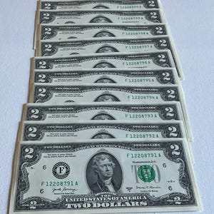May include: A collection of ten United States two-dollar bills arranged in a vertical overlapping stack. Each green and black banknote features a portrait of Thomas Jefferson, the text Two Dollars, and sequential serial numbers starting with F12208791A.