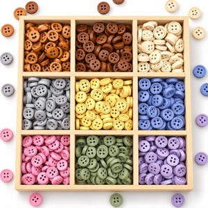 May include: A wooden organiser box containing nine compartments filled with round, four-hole buttons in various colours including brown, beige, grey, yellow, blue, pink, green, and purple, with additional loose buttons scattered around the box.