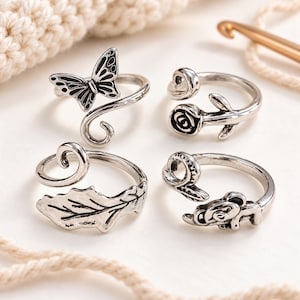 Pode incluir: Um conjunto de quatro anéis de metal prateado com bandas abertas e ajustáveis. Os designs incluem uma borboleta, uma rosa, uma folha texturizada e uma pequena figura animal, com detalhes oxidados escuros para realçar os padrões.