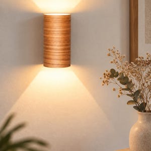 May include: A cylindrical wall sconce featuring a light brown wood grain finish. The fixture emits a warm, bright glow from both the top and bottom openings, casting distinct triangular beams of light onto the wall surface above and below it.