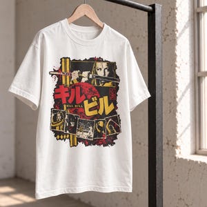 May include: A white short-sleeve t-shirt featuring a graphic print of the film Kill Bill. The design includes a large red sun, Japanese text, and multiple film stills showing characters with swords and a yellow jumpsuit against a black background.