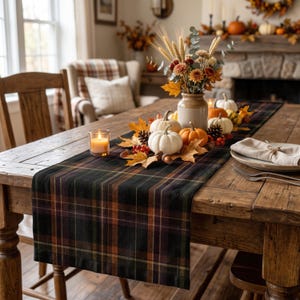 May include: A rectangular table runner featuring a dark plaid pattern in shades of forest green, deep orange, and black. The runner is laid across a wooden table and decorated with white pumpkins, orange pumpkins, pinecones, and autumn leaves.