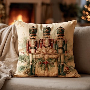 May include: A square throw pillow featuring a printed illustration of three nutcracker figures standing behind a large gift box tied with a ribbon. The design includes green pine branches, red berries, and a cream-colored background.