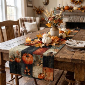Op de afbeelding: Een rechthoekige tafelloper met een patchworkontwerp van oranje pompoenen, herfstbladeren en tekst op een donkere achtergrond. De loper ligt op een houten tafel met een middelpunt van witte pompoenen, tarwe en bloemen.