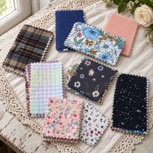 May include: A collection of rectangular fabric swatches with pinked edges, featuring various patterns including plaid, floral prints in blue, black, and peach, a light purple gingham check, a solid navy blue, and a dark navy with white speckles.