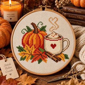 Op de afbeelding: Een ronde houten borduurring met een kruissteekpatroon van een oranje pompoen, een witte mok met een rood hart, stoom en kleurrijke herfstbladeren op een crèmekleurige stof, wat een seizoensgebonden ontwerp vormt.