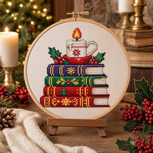 May include: A circular wooden embroidery hoop containing a cross-stitch pattern of a stack of four books topped with a lit candle in a mug. The books are purple, green, red, and red, decorated with snowflakes and holly. The candle has a yellow flame.