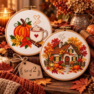 Può includere: Due telai da ricamo circolari in legno contenenti arte a punto croce. Uno presenta una zucca, foglie autunnali e una tazza con un cuore. L'altro raffigura un cottage circondato da fogliame autunnale, zucche e una lanterna. Un'etichetta riporta Set of 2.