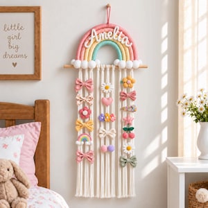 May include: A wall-hanging macrame hair clip organizer featuring a rainbow arch with the name Amelia in white script. The rainbow has pink, yellow, and teal bands with white pom-pom clouds. Multiple white cords hang down, displaying various fabric hair bows, flowers, hearts, and fruit-shaped clips.