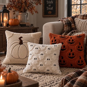 Peut inclure: Trois coussins décoratifs carrés en tissu texturé. Un coussin crème présente une citrouille brodée, un coussin blanc arbore un motif répétitif de petits fantômes blancs, et un coussin orange affiche un motif répétitif de citrouilles d'Halloween.