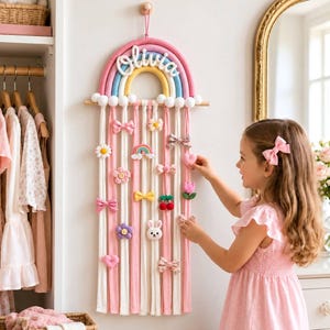 May include: A wall-hanging rainbow organizer featuring the name Olivia in white script. The rainbow has pink, blue, and yellow arches with white pom-poms. Multiple vertical fabric strips in pink and white hold various hair clips and bows.