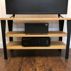 May include: A black TV stand with two shelves made of light wood. The stand has black metal legs and is holding a black TV, a black speaker, and a black receiver.