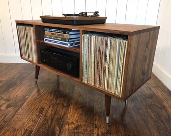 May include: A walnut wood record player stand with a turntable on top, a shelf for an amplifier, and two shelves for vinyl records. The stand has four tapered legs.