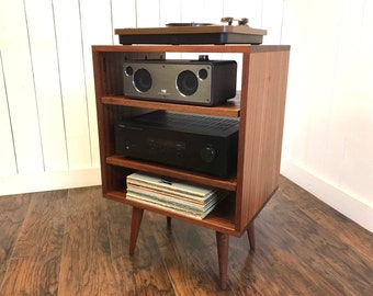 May include: A wooden record player stand with three shelves. The top shelf has a turntable, the middle shelf has a stereo receiver, and the bottom shelf has a stack of vinyl records. The stand has four legs.