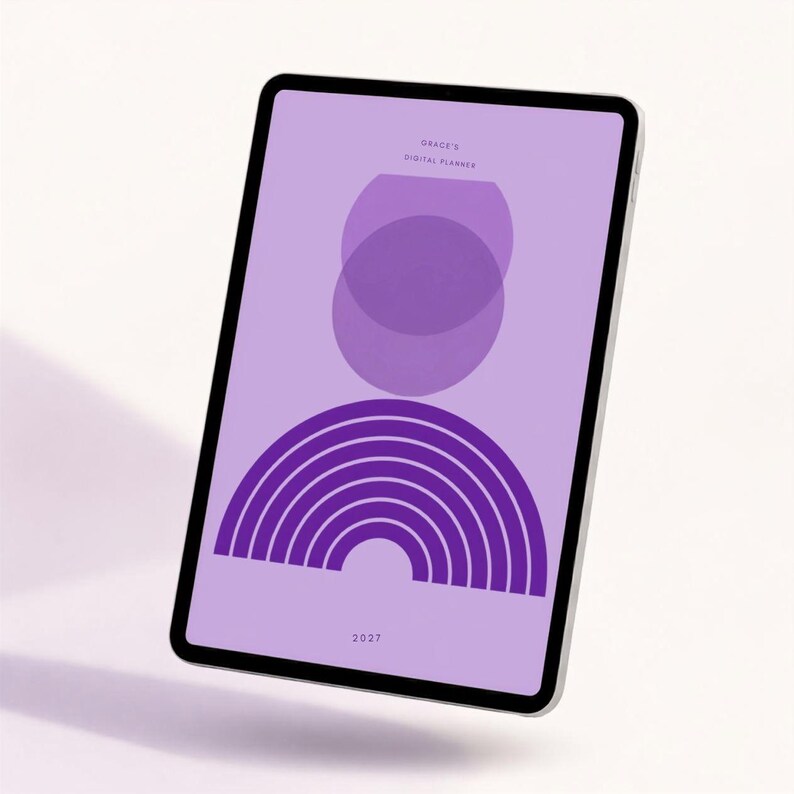 Pode incluir: Um tablet exibindo um planejador digital com fundo roxo claro. A tela apresenta o texto Grace's Digital Planner e 2027, juntamente com um design gr&aacute;fico composto por dois c&iacute;rculos roxos sobrepostos e um arco-&iacute;ris semicircular roxo escuro.