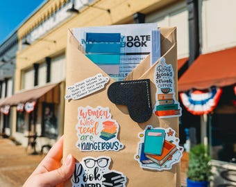 May include: A brown paper envelope containing a date book, blue sticky tabs, and a pen. The exterior is decorated with stickers featuring book-themed illustrations, glasses, and text including "Women who read are dangerous" and "Book Nerd."