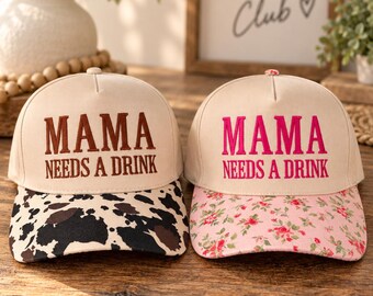 May include: Two beige baseball caps with the embroidered text MAMA NEEDS A DRINK. The left cap features a brown cow print brim, while the right cap has a pink floral pattern on the brim. Both caps have a structured crown and a button on top.