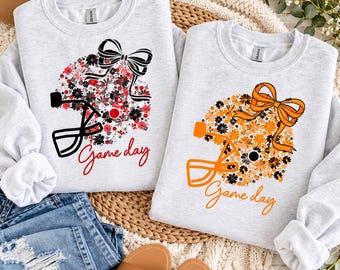 May include: Two light grey crewneck sweatshirts featuring a football helmet graphic filled with floral patterns and a bow. One helmet is red, black, and white with the text "Game day" in red. The other is orange, black, and white with "Game day" in orange.