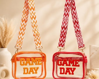 May include: Two clear rectangular crossbody bags with orange and red trim. Each bag features the text GAME DAY in fuzzy block letters. The bags have detachable woven shoulder straps with orange and red chevron and diamond patterns respectively.