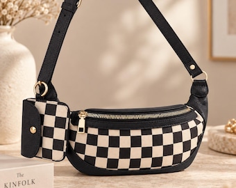 May include: A crescent-shaped crossbody bag featuring a black and white chequered pattern with black leather-like trim, a gold-tone zip, an adjustable strap, and a small detachable matching chequered pouch attached to the side ring.