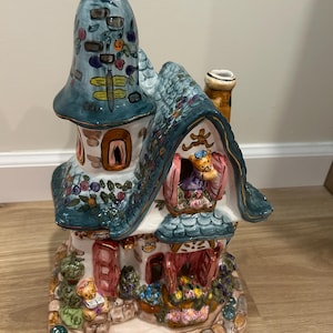 May include: A ceramic house figurine featuring a tall blue turret, a shingled roof, and painted floral details. The structure includes small teddy bear figures in the windows and at the entrance, with a stone-textured base and a chimney.