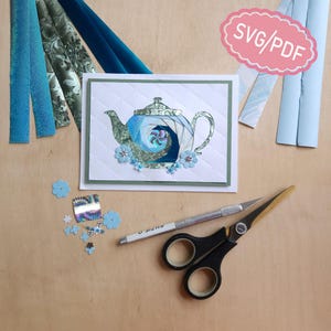 May include: A greeting card featuring a paper-pieced teapot design in shades of blue and green, with a pink scalloped label reading SVG/PDF. The card is surrounded by paper strips, small blue flower cutouts, a craft knife, and black scissors.