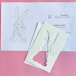 May include: A papercraft pattern featuring a bunny design, with numbered sections for assembly. The pattern is accompanied by a cut-out bunny shape. The text "SuzieQ Papercrafts" and "Pattern #107" are visible on the paper.