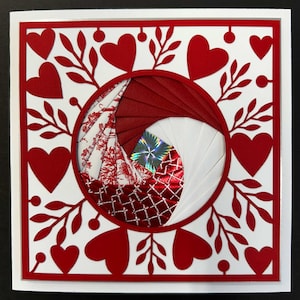 May include: A square, layered paper art piece with a Valentine's Day theme. The design features a red and white color scheme, with red hearts and leaf cutouts surrounding a circular center with folded paper and a holographic element.