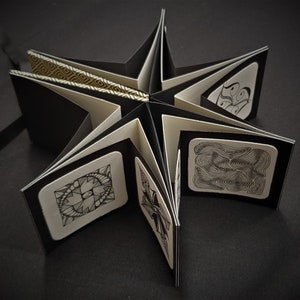 May include: A star-shaped, handmade art book with black covers and white pages. Each page features a unique black and white abstract drawing. The book is open, revealing the intricate designs and patterns within.